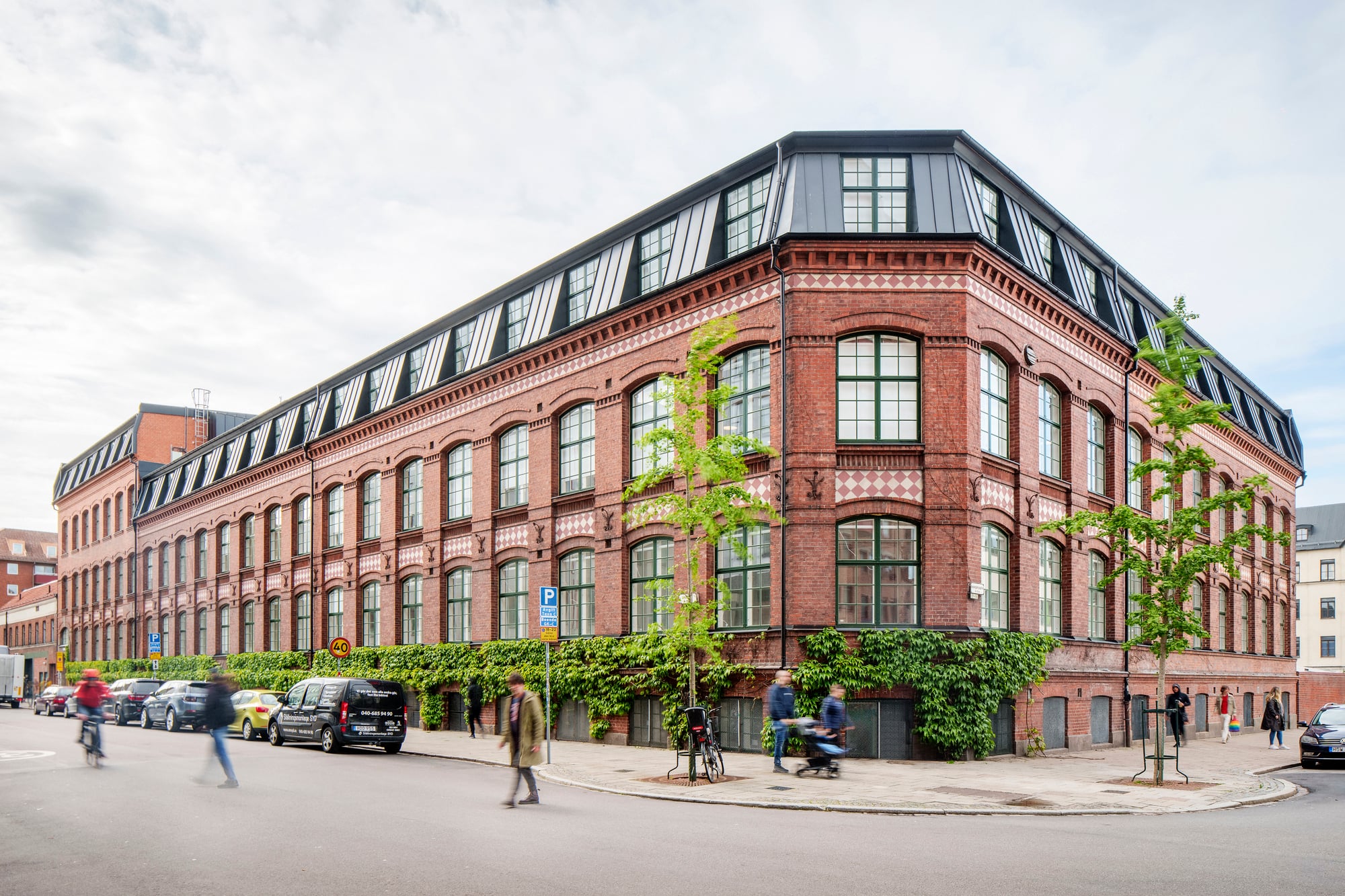 Ett stort hörnhus i rött tegel med stora spröjsade fönster och svart takvåning, omgivet av träd, parkerade bilar och förbipasserande cyklister och fotgängare i en stadsmiljö.
