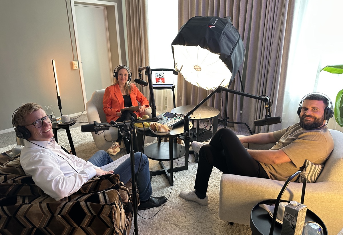 Tre personer spelar in en podd i ett vardagsrumsliknande rum. De sitter i varsin fåtölj runt ett litet bord med mikrofoner, hörlurar och inspelningsutrustning, upplysta av en stor studiobelysning framför gardiner och ett fönster.