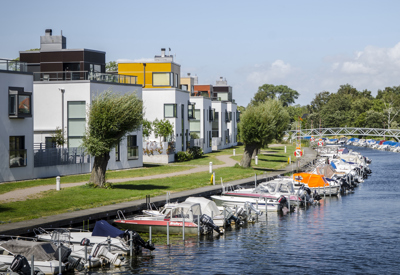 En rad moderna, vita bostadshus med platta tak belägna längs en kanal. Framför byggnaderna finns en gångväg kantad av träd och gräs. Flera små båtar är förtöjda längs vattenkanten.
