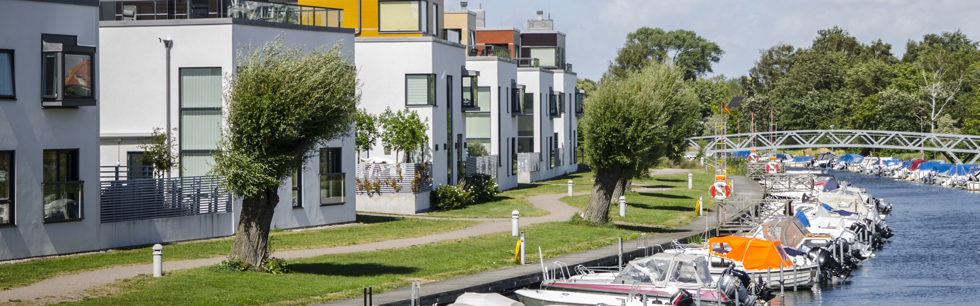 En rad moderna, vita bostadshus med platta tak belägna längs en kanal. Framför byggnaderna finns en gångväg kantad av träd och gräs. Flera små båtar är förtöjda längs vattenkanten.