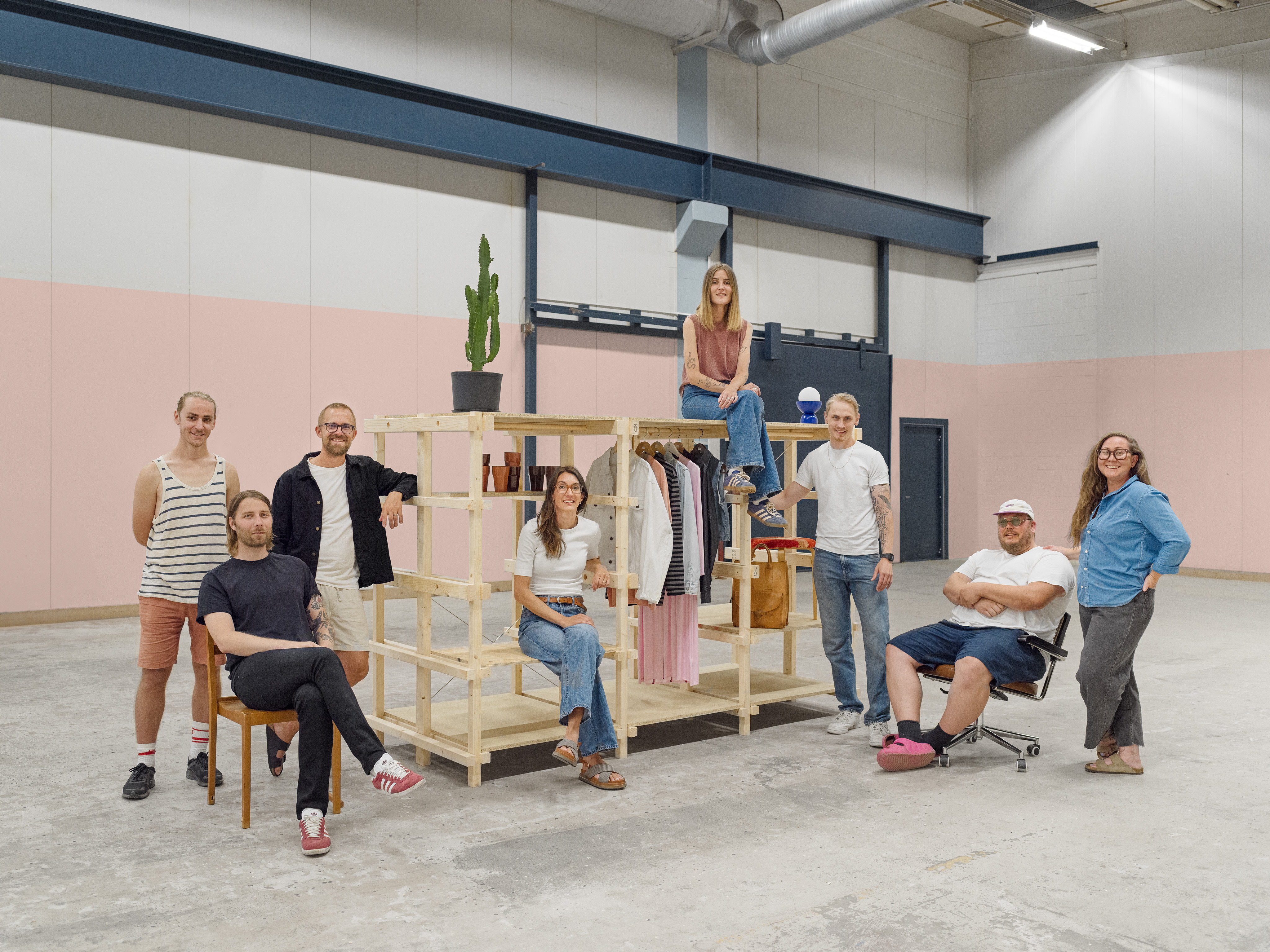 En grupp på åtta personer poserar i ett stort, ljust industriliknande rum. De står och sitter runt en hylla i ljust trä där kläder och några inredningsdetaljer hänger och står. En person sitter ovanpå hyllan, två sitter på stolar framför, och resten står bredvid. Väggarna är målade i rosa och grått, och golvet är av betong.