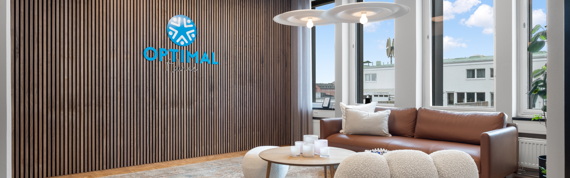 En modern loungedel på ett kontor med trägolv, en brun lädersoffa vid stora fönster, två vita fåtöljer och ett runt soffbord på en matta, framför en vägg med träpanel där företagets logotyp Optimal syns
