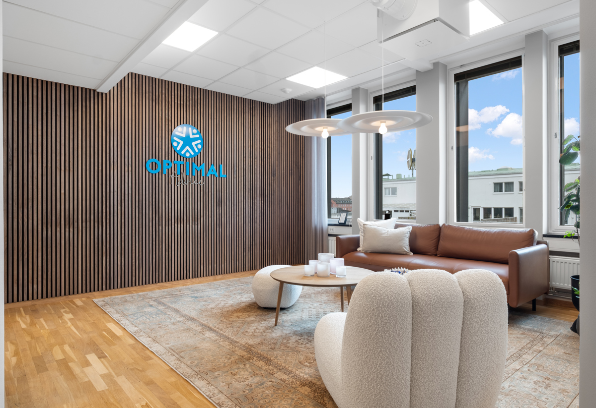 En modern loungedel på ett kontor med trägolv, en brun lädersoffa vid stora fönster, två vita fåtöljer och ett runt soffbord på en matta, framför en vägg med träpanel där företagets logotyp Optimal syns
