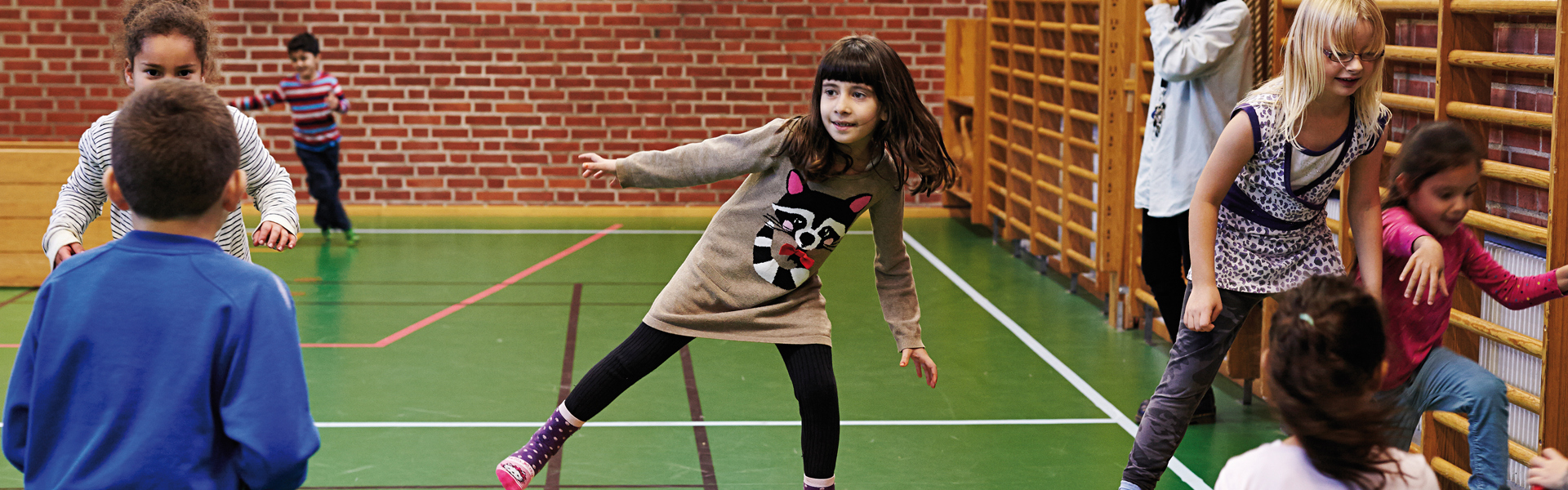 Ett gäng barn leker och tränar balans i en gymnastiksal. Ett barn går balanserande på en plint, medan andra barn tittar på eller väntar på sin tur. Gymnastiksalen har grönt golv, röda tegelväggar och ribbstolar längs väggarna.
