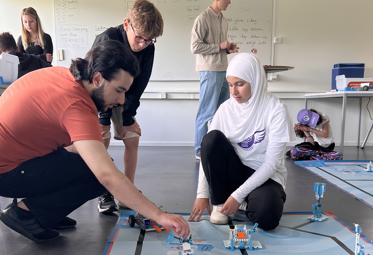 En grupp människor deltar i en robotskola där de bygger och programmerar robotar. En person i orange tröja och en i vit hijab fokuserar på en robot på golvet. Andra deltagare syns i bakgrunden, engagerade i olika aktiviteter.