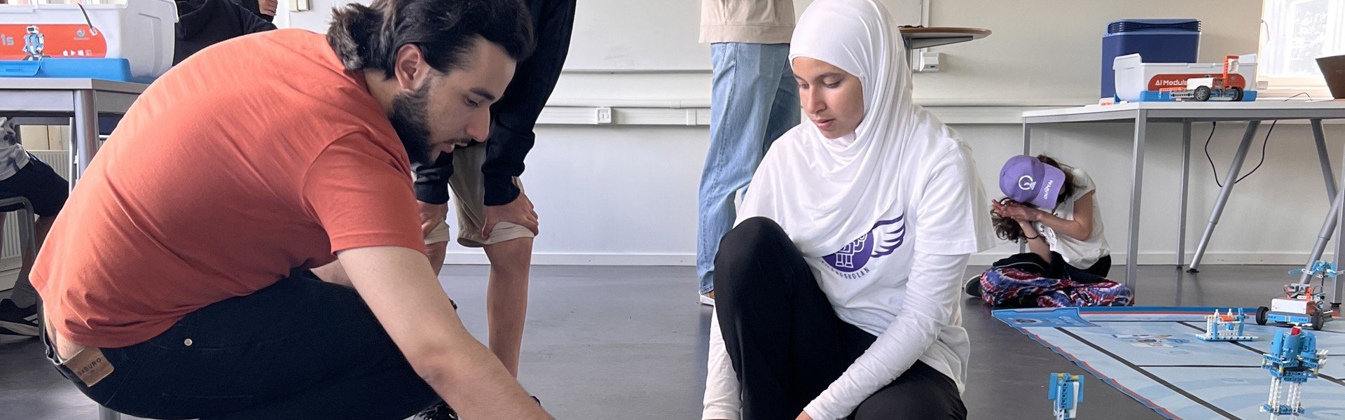 En grupp människor deltar i en robotskola där de bygger och programmerar robotar. En person i orange tröja och en i vit hijab fokuserar på en robot på golvet. Andra deltagare syns i bakgrunden, engagerade i olika aktiviteter.
