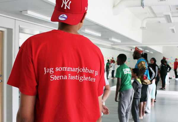 En person i röd tröja och keps står i ett rum med spegelväggar. På tröjan står det "Jag sommarjobbar på Stena Fastigheter." Flera personer, inklusive barn och vuxna, står på rad och deltar i en aktivitet.