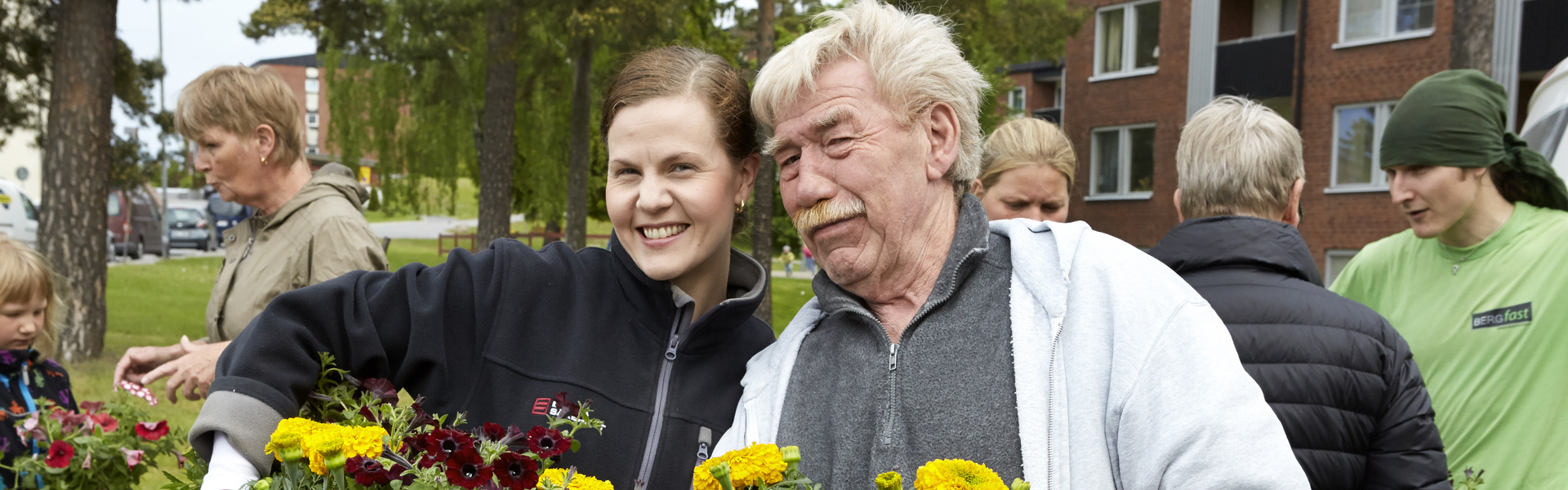 En grupp människor utomhus som håller i blomkrukor med färgglada blommor. En man och en kvinna står i förgrunden, båda leende, med fler personer i bakgrunden. Byggnader och träd syns i bakgrunden.