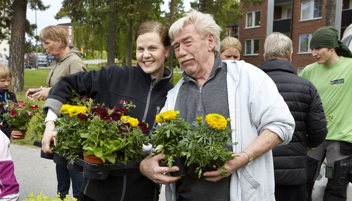 En grupp människor utomhus som håller i blomkrukor med färgglada blommor. En man och en kvinna står i förgrunden, båda leende, med fler personer i bakgrunden. Byggnader och träd syns i bakgrunden.