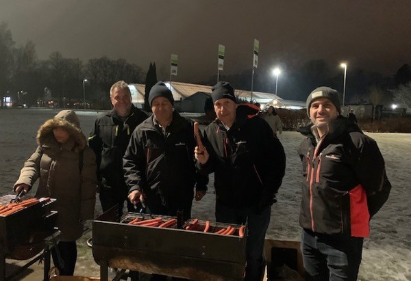Fem personer står vid en grill utomhus under en vinterkväll och grillar korv.
