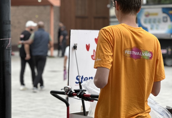 Ung person som står med ryggen mot på ett torg i en orange tröja där det står festivalvärd.