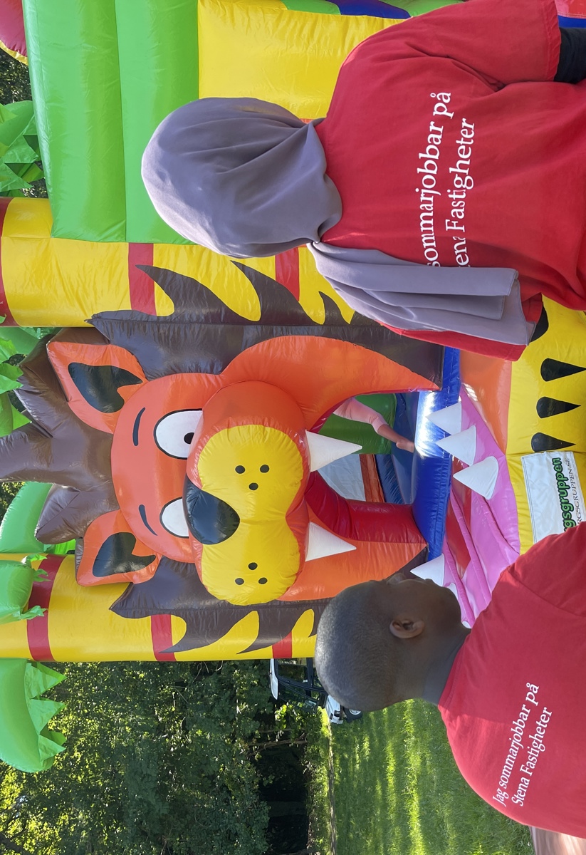 Hoppborg med lejon och palmer. I förgrund två ungdomar med röda sommarjobbs-tröjor på.