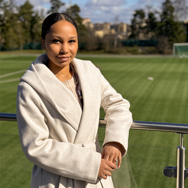 Porträtt av ung kvinna som står lutad mot räcke med fotbollsplan i  bakgrund.