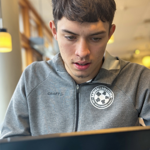 Porträtt av ung kille som sitter framför en skärm. Killen har grå tröja med Samhällsmatchen-logga.