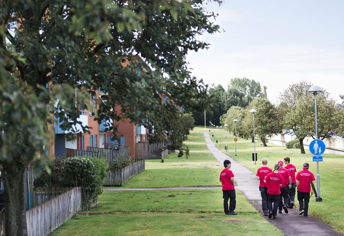 En grupp unga människor med röda tröjor där det står "sommarjobbare" på texten som går tillsammans i en grupp i ett grönt och frodigt kvartert med en gångstig och mycket gräsmatta