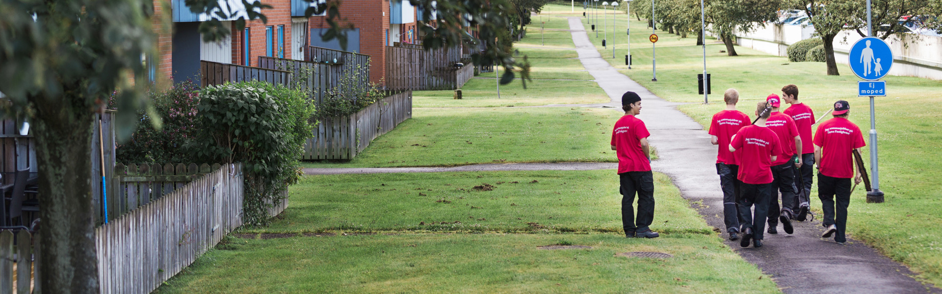 En grupp unga människor med röda tröjor där det står "sommarjobbare" på texten som går tillsammans i en grupp i ett grönt och frodigt kvartert med en gångstig och mycket gräsmatta
