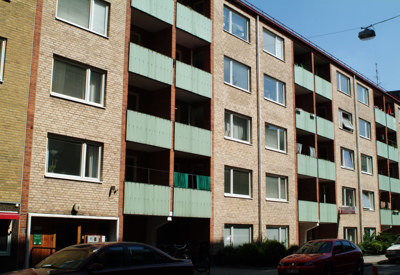 En flerfamiljsbyggnad med en tegelstensfasad och gröna balkonger. Byggnaden har fem våningar, var och en med flera fönster och balkonger. Bilar är parkerade framför byggnaden på gatan, och en cykel är synlig nära ingången. Himlen är klar, vilket indikerar bra väder.