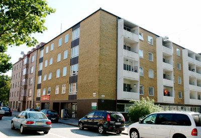 En flerfamiljsbyggnad med en gul tegelstensfasad och flera balkonger. Flera bilar är parkerade längs gatan framför byggnaden. Scenen verkar vara i ett urbant bostadsområde med träd som kantar gatan.