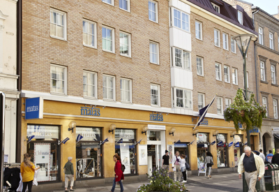 Folk promenerar på en solig gågata framför butiken Matas, som ligger i en ljus tegelbyggnad med flera våningar och ett mörkbrunt tak. Skyltar och fönster visar butikens namn, och en stor blomkruka står på trottoaren.