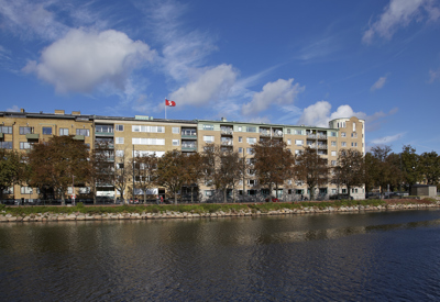 Ett flerbostadshus i ljusgul tegel med fem våningar, beläget intill en kanal. Träd står i rad framför byggnaden och en röd Stena flagga vajar från taket. Himlen är blå med några vita moln.