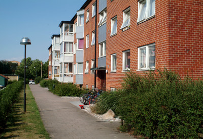 Ett rött trevånings flerfamiljshus med vita fönster och entrébalkonger i olika färger. Framför huset löper en gångväg kantad av häckar, cyklar står parkerade vid entrén och det är soligt med klarblå himmel, vilket ger området en trivsam och välskött känsla.