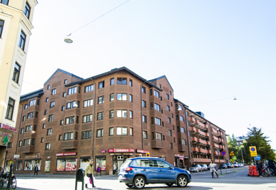 En femvånings hörnbyggnad i rött tegel med butikslokaler i markplan och bostäder ovanför. Byggnaden har ett brutet tak och flera fönster i regelbunden rytm. På gatuplanet finns skyltar för bland annat en restaurang och en livsmedelsbutik. En blå bil kör förbi i korsningen, och flera fotgängare syns på trottoarerna i det soliga vädret.