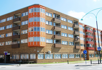 Ett fyravåningshus i gult tegel med orange hörnburspråk och mörkbruna balkonger. Byggnaden ligger vid en gata med övergångsställe och blå gatubelysning.