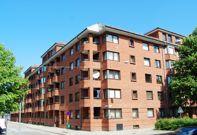 Ett större bostadskomplex i rödbrunt tegel, med fem våningar och ett hörnläge som skapar en tydlig struktur i stadsbilden. Byggnaden har enkla balkonger, varierande fönsterstorlekar och inslag av takkupor som bryter upp volymen. Grönskan runt huset bidrar till en trivsam miljö.