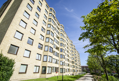 Ljust flerfamiljshus i tegel med många våningar, sett från marknivå en solig dag. Gröna träd och en gångväg löper längs byggnaden.