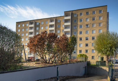 Ett gult flerbostadshus i åtta våningar med ljusa balkonger längs ena sidan. Framför byggnaden finns en liten lekplats och flera träd, varav några med rödbruna höstlöv. I förgrunden syns en sluttande gångväg kantad av buskar, som leder upp mot huset. Soligt väder med klarblå himmel bidrar till en ljus och trivsam känsla.