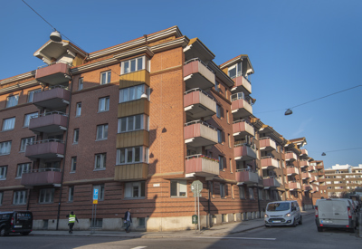 Ett större rödteglat bostadshus med flera balkonger, beläget vid en gata med parkerade bilar och några gående personer. Solen lyser mot byggnaden från vänster och himlen är klarblå.