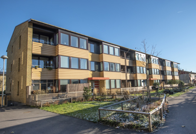 Lågt flerbostadshus i tre våningar med fasad i gult tegel och träpanel. Byggnaden har balkonger och stora fönster, och framför huset finns en gräsmatta med inslag av snö, buskar och en gångväg. Klarblå himmel och soligt väder.