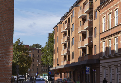 Stadsgata kantad av rödbruna flerbostadshus med balkonger, parkerade bilar och människor i rörelse. Träd skymtar mellan husen och himlen är delvis molnig i bakgrunden.
