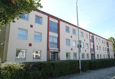 En trevånings flerfamiljsbyggnad med ljus fasad, röda detaljer kring fönster och entréer samt gröna buskar som löper längs hela byggnadens framsida. En trottoar och en gatlykta syns i förgrunden under en klarblå himmel.