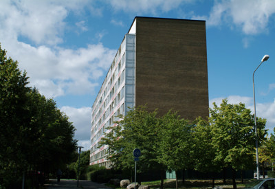 Ett högt flerfamiljshus med mörk gavelfasad och ljusa, rutmönstrade balkonger längs långsidan. Byggnaden är omgiven av gröna träd och gångvägar, med en klarblå himmel och moln i bakgrunden. En gatlykta och ett cykelskylt syns i förgrunden.