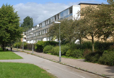 En lång, vit flerbostadsbyggnad i tre våningar med balkonger och stora fönster. Byggnaden är omgiven av grönska, träd och buskar, med en gång- och cykelväg som löper längs framsidan. En person promenerar i fjärran under en delvis molnig himmel.