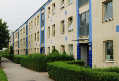 En lång, ljusgul trevånings flerbostadsbyggnad med blå detaljer kring fönster och entréer. Byggnaden kantas av välklippta häckar och en gångväg som löper längs med fasaden. Träd och grönska omger området under en mulen himmel.