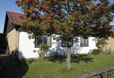 Ett enplanshus med ljus fasad, tegelväggar och rött tegeltak. Framför huset står ett rönnbärsträd med röda bär som lyser upp trädgården. Gräsmattan är välskött och det finns en smal gångväg längs sidan av huset där en cykel står parkerad. Soligt väder med blå himmel.