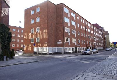 Ett hörn av ett kvarter med rödteglade flerbostadshus i fyra våningar. Fönster i regelbundet mönster täcker fasaden. Några bilar är parkerade längs gatan och trottoaren är kantad av stenplattor och gatsten. Solen kastar skuggor mot husväggen.