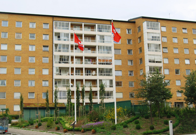 Ett flerbostadshus i gult tegel med sju våningar och flera inglasade balkonger. Framför byggnaden finns en välskött trädgård med buskar, träd och två flaggstänger med röda Stena flaggor. Ljuset är mjukt och himlen mulen, vilket skapar ett stillsamt intryck av miljön.