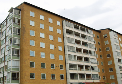 Ett större bostadshus i gult tegel med flera våningar och inglasade balkonger. Byggnaden har en funktionell och enkel stil, med fönster i symmetriska rader. Himlen är molntäckt och ljuset är jämnt, vilket ger ett neutralt intryck av miljön.
