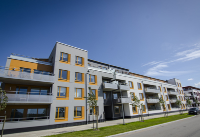 Ett modernt flerfamiljshus med tre till fyra våningar. Fasaden är ljusgrå och vit med orange detaljer runt fönstren, vilket ger byggnaden ett fräscht och samtida utseende. Huset har stora glasade balkonger och ligger längs en bred gata med små nyplanterade träd och grön gräsmatta. Soligt väder med blå himmel.