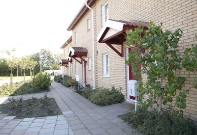 En rad med moderna radhus i två våningar byggda i gul tegel. Varje entré har ett rött dörrparti med ett litet vitt tak över. Framför husen finns en smal trädgårdsremsa med buskar och mindre växter, och en stenlagd gångväg löper längs hela raden. Soligt väder med klar himmel.
