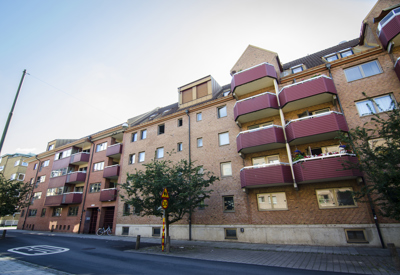 Flerbostadshus i rödbrunt tegel med flera våningar och balkonger med vinröda räcken mot en lugn gata. Träd står planterade längs trottoaren, och en skylt för gångtrafik syns framför ett övergångsställe. Himlen är klar med ett svagt ljus.