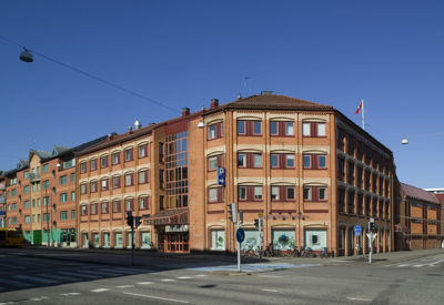 En större hörnbyggnad i rödtegel med fyra våningar och sadeltak. Byggnaden har rundade hörn, många fönster och en glasad entrédel i mitten. På trottoaren framför huset står cyklar parkerade och ett par trafikskyltar syns. Gatan framför är bred med övergångsställen och trafikljus, och himlen är klarblå.
