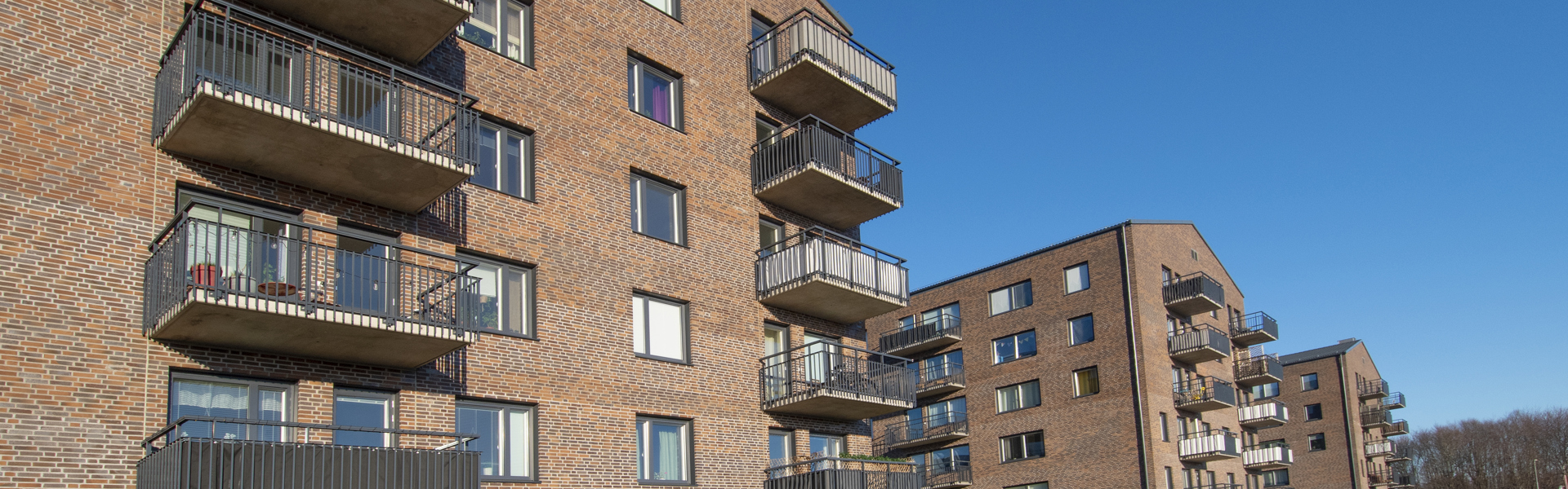 Flerbostadshus i rött tegel med balkonger, omgivna av grönområden. Byggnaderna står mot en klarblå himmel, vilket ger en tydlig kontrast.