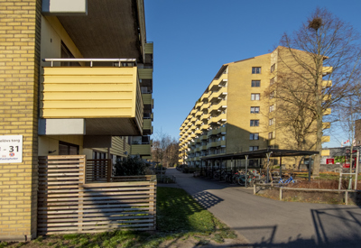 Lång gångväg mellan två flerbostadshus i gult tegel. Till vänster finns en skylt med adresserna "Nöbbelövs torg 29–31". Flera balkonger syns längs fasaderna, och det finns cykelställ på höger sida. Träden är kala, och solen skiner från en klarblå himmel.