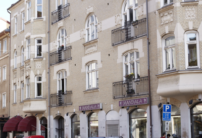 En vacker stadsgata med en ljus putsad sekelskiftesbyggnad i flera våningar. Fasaden är dekorerad med ornament och smidesbalkonger, och ett hörntorn med spira reser sig mot himlen. Längs trottoaren står parkerade bilar och en butik har skyltfönster i bottenvåningen.