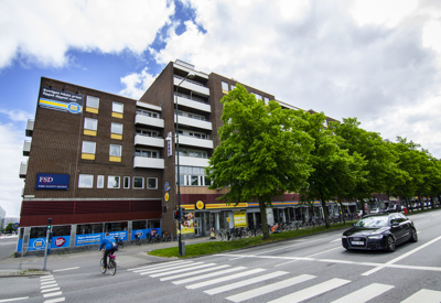 Ett större kvartershus i mörkbrunt tegel, med inslag av vita och glasade balkonger som skapar kontrast. Den ligger vid en större gata med tydlig stadskaraktär, där bottenvåningen rymmer butiker och service, vilket ger liv åt gatumiljön. Trädallén framför huset bidrar till en grön, inbjudande stadsbild.