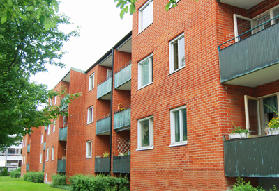 En röd tegelbyggnad med tre våningar och gröna balkonger, omgiven av grönska. Längs husets ena sida löper en gräsmatta och träd som skapar en skuggig, trivsam miljö. Växtlighet och balkongplanteringar ger ett hemtrevligt intryck i bostadsområdet.